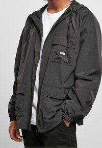 Black lightweight jacket with a hood, zip closure, two front pockets, and elastic cuffs. Features a small logo patch on the chest pocket.