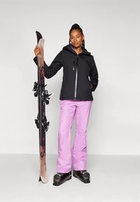 Ženska v črnem jopiču, roza smučarskih hlačah in smučarskih čevljih drži par smuči pokonci pred enobarvno sivo ozadjem.