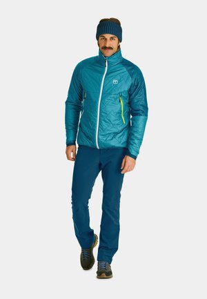 Teal insulated jacket with a high collar and front zip. Features a yellow zippered pocket and paired with blue pants and hiking shoes.