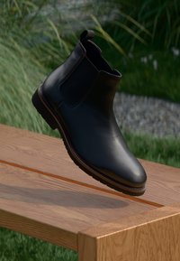 Bottine Chelsea en cuir noir au design épuré, avec des panneaux latéraux élastiques, un bout rond et une semelle marron. Présentée sur une surface en bois.