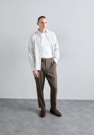 Chemise à boutons légèrement rayée en blanc et gris, associée à un pantalon tailleur marron et des chaussures en cuir marron. Coupe simple et moderne.