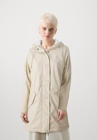 Beige vanntett jakke med snorhette, trykknapper, to frontlommer og en myk hvit fôr. Glatt tekstur, midtlengde design.