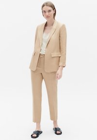 Beige linnen pak met een op maat gemaakte blazer en slim fit broek, gecombineerd met een licht topje. Het model draagt zwarte slippers. Eenvoudig, strak ontwerp.
