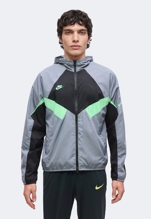 Νέος άνδρας που φοράει γκρι αδιάβροχο Nike με μαύρες και πράσινες λεπτομέρειες και μαύρο παντελόνι με πράσινο λογότυπο Nike, στέκεται μπροστά σε απλό φόντο.