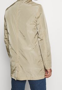 Beige lättviktsjacka med en slät, blank yta. Har rak snitt, sidofickor och minimalistisk design.