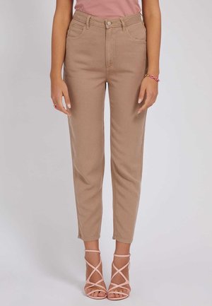 Kobieta ubrana w beżowe spodnie z wysokim stanem, przednimi kieszeniami i zapięciem na guziki, w połączeniu z różowymi sandałami na wysokim obcasie z paskami.