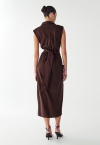 Robe midi sans manches marron avec un col et une taille nouée, fabriquée dans un matériau doux. Texture lisse avec un design simple, vue de dos.