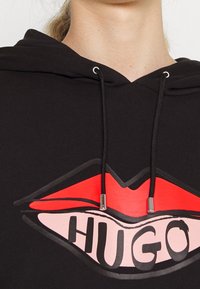 Černá mikina s kapucí a šňůrkami, která má výraznou grafiku červených rtů a slovo "HUGO" v tučném černém písmu. Měkká, texturovaná tkanina.