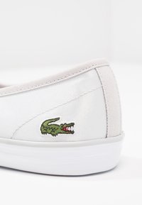 Chaussure blanche à enfiler en toile avec un emblème de crocodile vert. La semelle est blanche avec un bord texturé, et la tige a une finition lisse.