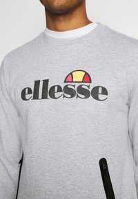 Primer plano de una persona que lleva un sudadera gris claro con el logotipo negro "ellesse" y un semicírculo colorido encima del texto, con bolsillos con cremallera negros.