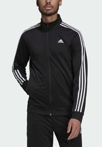 Chaqueta de pista negra hecha de una tela suave con un alto cuello, acento de tres rayas blancas en las mangas y un pequeño logo en el pecho.