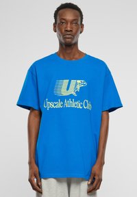 Mėlynas medvilnės marškinėlis su dideliu geltonu piešiniu, kuriame užrašyta "Upscale Athletic Club" su stilizuotu "U" ir varlės iliustracija.