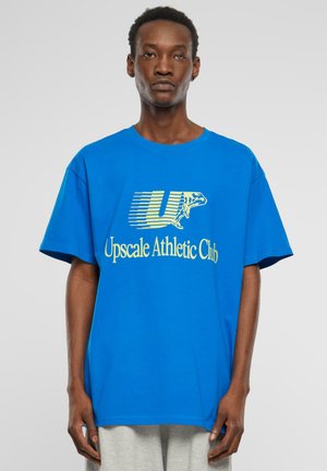 Blå bomulds t-shirt med en stor gul grafisk print, der står "Upscale Athletic Club" med et stiliseret "U" og en frø-illustration.