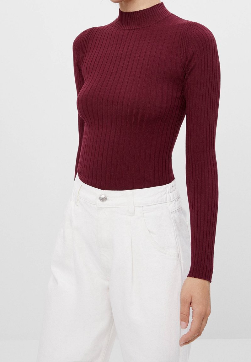 Burgundy ribbad polokragsweater med långa ärmar, åtsittande design, kombinerad med vita byxor med hög midja som har knäppning.