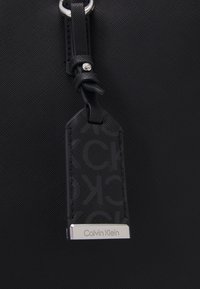 Czarna torba tote z teksturowanej tkaniny z srebrnym pierścieniem i czarną skórzaną zawieszką z monogramem. Zawieszka pokazuje logo "Calvin Klein".