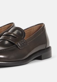 Smeđe kožne mokasinke s poliranim završetkom, ukrašene trakicom i niskom crnom petom. Glatka tekstura s detaljnim šavovima.