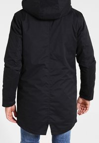 Homme portant un manteau d'hiver noir à capuche, vu de dos, avec un jean bleu visible sous le manteau.