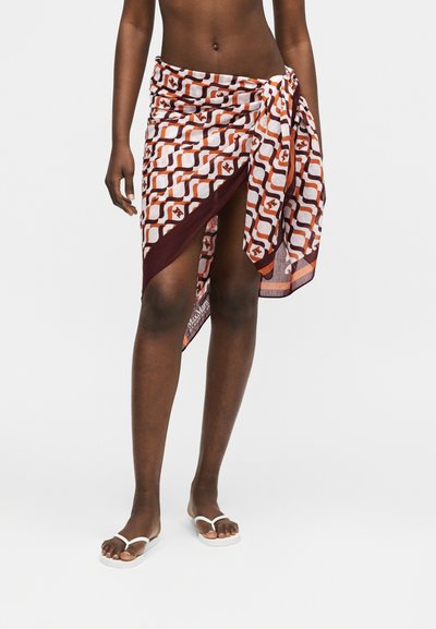 Personne à la peau foncée portant une jupe portefeuille à motifs géométriques marron, orange et blanc, ainsi que des tongs blanches, debout sur un sol blanc.
