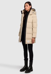 Woman wearing beige hooded puffer coat over black outfit with black ankle boots, standing against a plain light gray background.