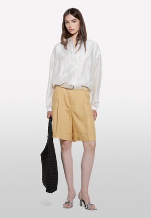 Donna con capelli lunghi e scuri che indossa una camicetta bianca larga, pantaloncini beige a vita alta, tacchi argento e tiene una borsa tote nera intrecciata.