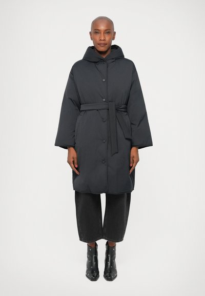 Black padded coat with a hood, buttons, and a tied belt. Features wide sleeves and a relaxed fit, worn over loose dark trousers and black ankle boots.