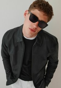 Schwarze Lederjacke mit Reißverschluss, glatter Textur und Steppdetails. Trägt sie über einem schwarzen Pullover und mit quadratische Sonnenbrille.