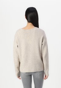 Beige ribbed sweater with long sleeves, deep rounded neckline, and relaxed fit. Soft texture. Worn with gray jeans.
