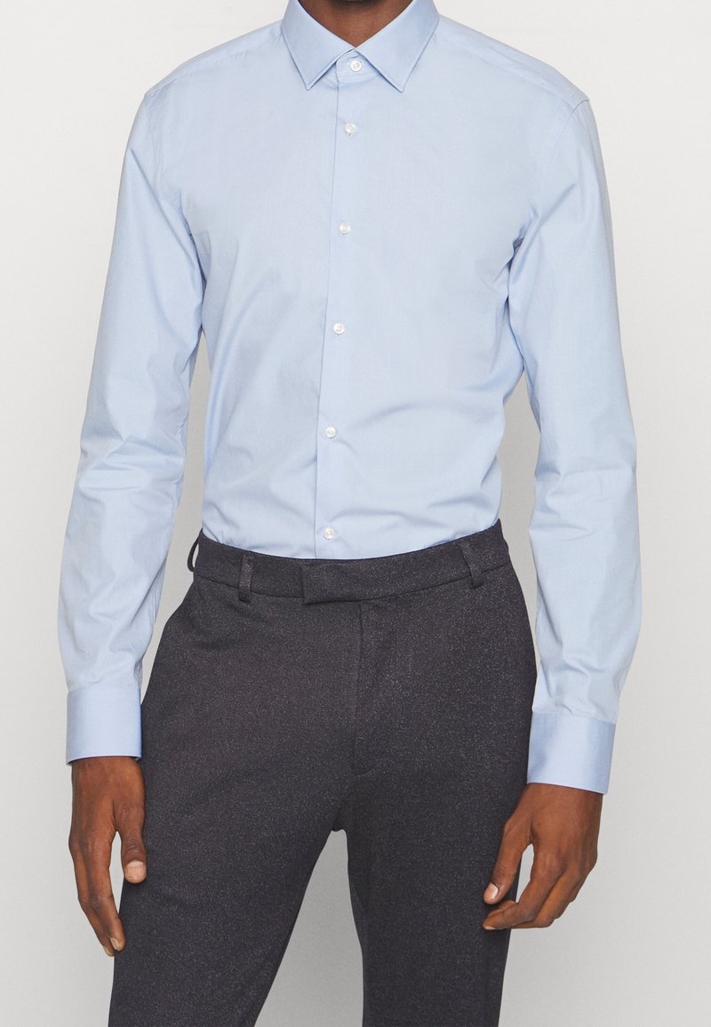 Homme portant une chemise boutonnée bleu clair à manches longues, rentrée dans un pantalon de costume gris foncé, sur un fond uni.