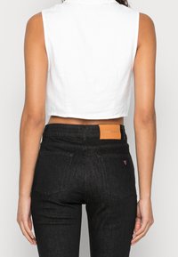 Croc top bianco senza maniche abbinato a jeans di denim neri, con un'etichetta beige sulla vita e un accento a forma di cuore viola sulla tasca posteriore.