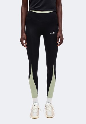 Person trägt schwarze Leggings mit hellgrünen Einsätzen an den Waden, ein schwarzes Top, weiße Socken und weiße Turnschuhe und steht aufrecht.