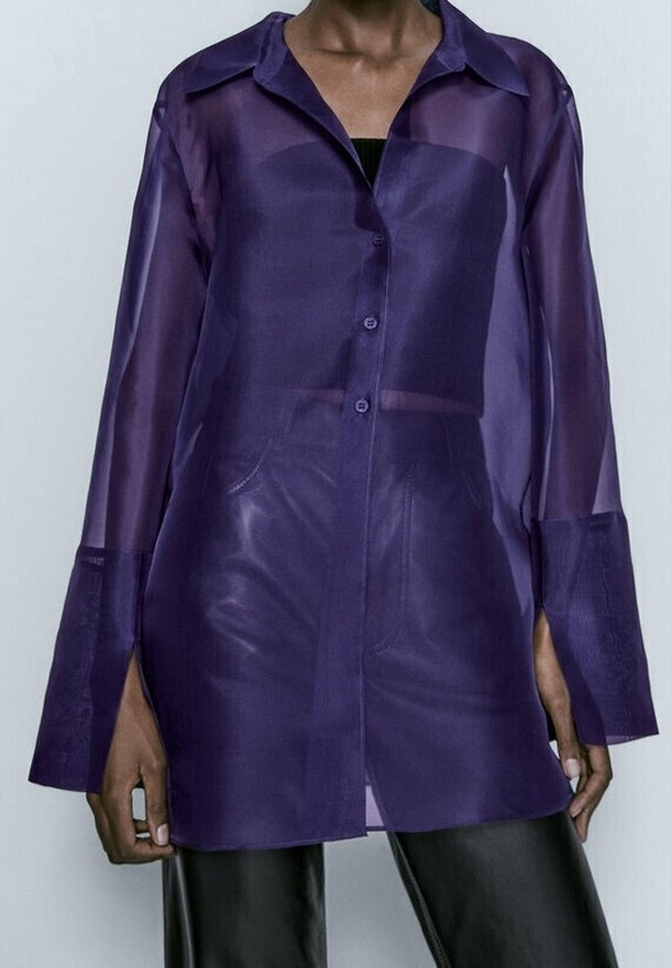 Personne portant une chemise violette longue à manches longues, boutonnée et transparente, par-dessus un haut noir et un pantalon noir, debout devant un fond clair uni.
