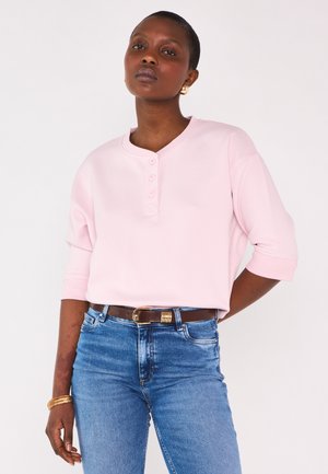 Donna con capelli corti che indossa una maglia rosa chiaro con bottoni, jeans blu, orecchini d'oro e braccialetti, in piedi contro uno sfondo bianco semplice.