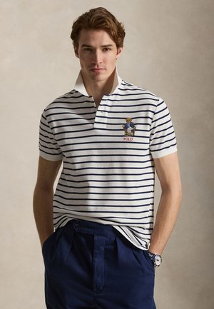 Mladi muškarac u bijeloj polo majici s mornarskim prugama i grbom medvjeda, tamnoplavim hlačama i ručnim satom, stoji ispred obične bež pozadine.