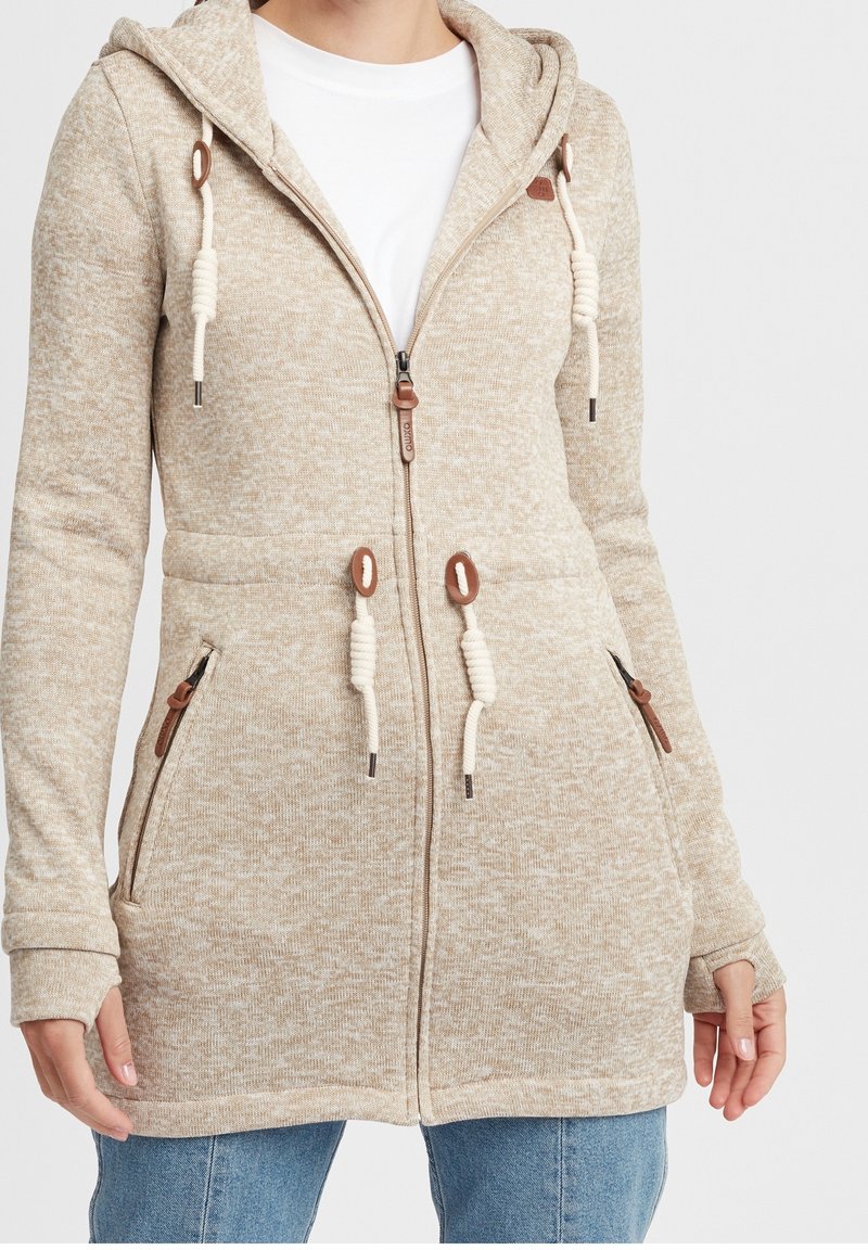 Sudadera con capucha beige con cremallera, hecha de tela texturizada. Cuenta con cordones y detalles de cuero, dos bolsillos laterales y puños acanalados.