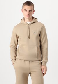 Felpa beige realizzata in tessuto morbido, con tasca a marsupio, cappuccio con coulisse e logo verde ricamato sul petto.