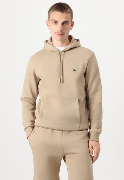 Hoodie beige en tissu doux, avec une poche kangourou, un capuchon à cordon, et un logo vert brodé sur la poitrine.