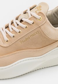 Baskets en daim beige avec lacets blancs, œillets dorés, semelle blanche épaisse et "Filling Pieces" embossé en or sur le côté.