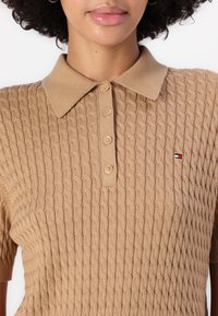 Polo shirt beige a maglia con un motivo a cavo testurizzato, quattro bottoni e un piccolo logo sul petto sinistro. Maniche corte.