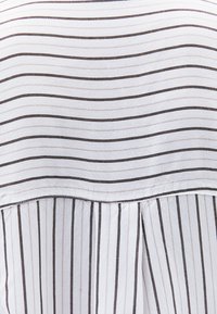 Top in tessuto bianco con sottili righe nere orizzontali e verticali, caratterizzato da un design con pieghe sul retro e una texture liscia.