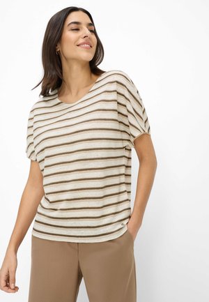 Kvinde iført en beige og brunstribet kortærmet top og lysebrune bukser, stående med den ene hånd i lommen mod en hvid baggrund.