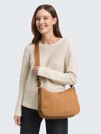 Tan suede shoulder bag with a textured finish, adjustable strap featuring engraved accents, paired with a cream long-sleeve shirt and dark jeans.