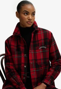 Red and black plaid jacket with a button-up front, collar, and a chest pocket featuring white embroidered brand logo. Warm fabric texture.