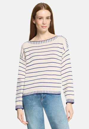 Frau, die einen weiß-blau gestreiften Pullover mit langen Ärmeln und hellblaue Jeans trägt, steht vor einem einfarbigen Hintergrund.