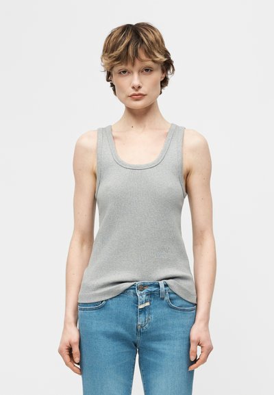 Jeune personne aux cheveux courts portant un haut sans manches gris et un jean bleu, debout devant un fond clair uni.