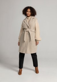 Beige wollen mengeling jas met een sjaalkraag, taille met riem en twee knopen; heeft geribde trui mouwen en is gecombineerd met zwarte broeken.