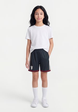 Ung person står med den ene hånd i lommen iført hvid t-shirt, sorte sportsshorts med emblem, hvide sokker og hvide sneakers.