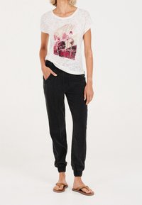 Vit kortärmad skjorta med grafiskt tryck, i kombination med svarta joggerbyxor med sidfickor och elastiska muddar. Bruna sandaler.