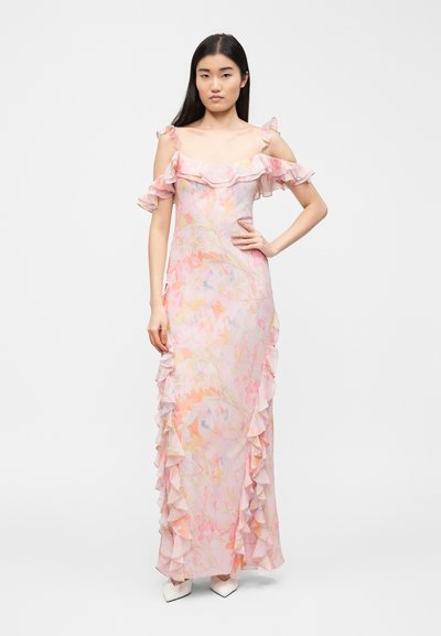 Γυναίκα με μακριά μαύρα μαλλιά που φοράει ροζ μακρύ φόρεμα με λουλούδια και ανοιχτούς ώμους με βολάν, στέκεται με το ένα χέρι στη μέση και λευκά μυτερά παπούτσια.