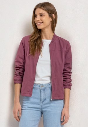 Jeune femme aux longs cheveux bruns portant une veste violette zippée, une chemise blanche et un jean bleu clair, debout contre un mur blanc texturé.