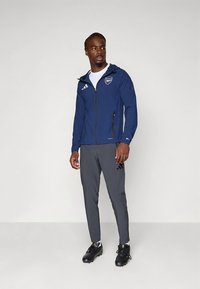 Marineblauer Windbreaker mit Kapuze und Reißverschluss, mit weißem Logo und drei Streifen. Graue Sporthose und schwarze Stollenschuhe. Einfaches Design, glatte Textur.